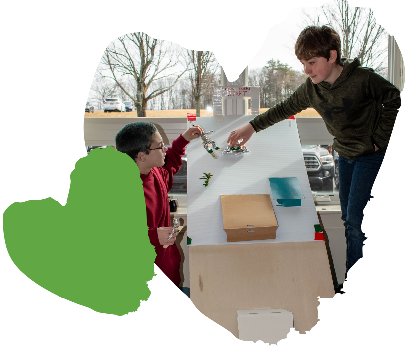 Two children are playing with a toy car on a white ramp indoors. One child is placing the car while the other watches. Trees and cars can be seen outside through the window. There is a graphic of a green heart to the left.