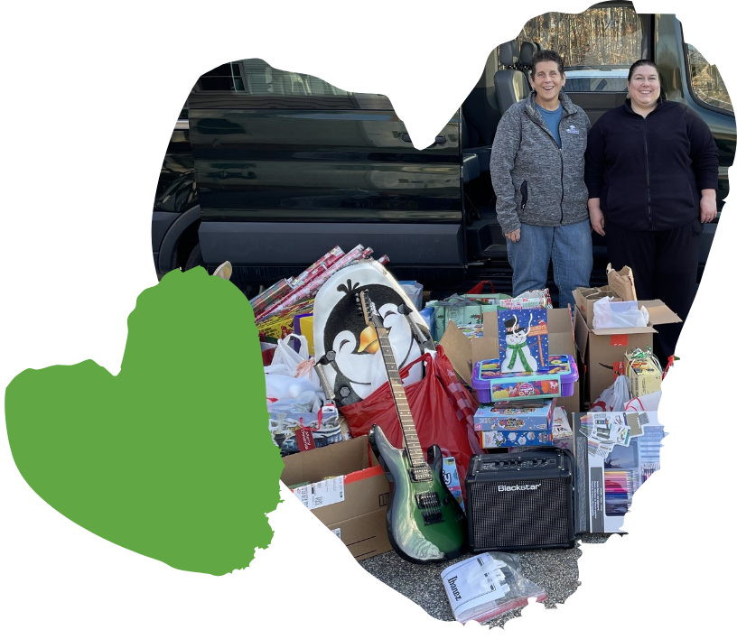 Two people stand next to a car filled with donations, including toys, a large stuffed penguin, a guitar, and an amplifier. Bags and boxes filled with various items are stacked on the ground in front of them. Graphic of a green heart is set to the left.