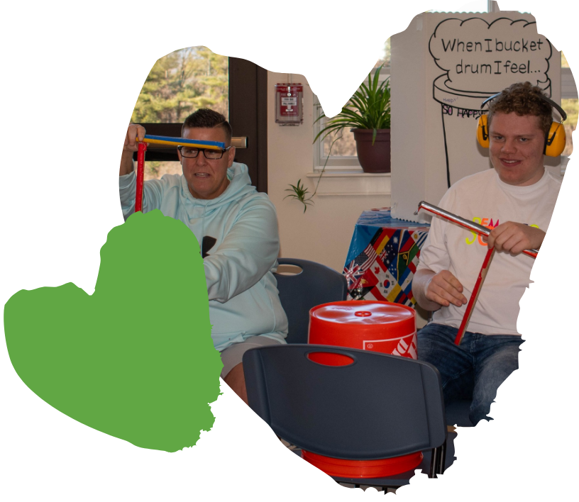 Two people wearing ear protection are drumming on plastic buckets in a campus room, creating a rhythm that echoes through the halls.