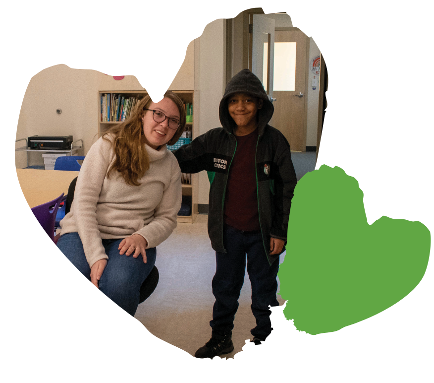 A woman seated next to a smiling child in a classroom, with a green heart outline overlay.