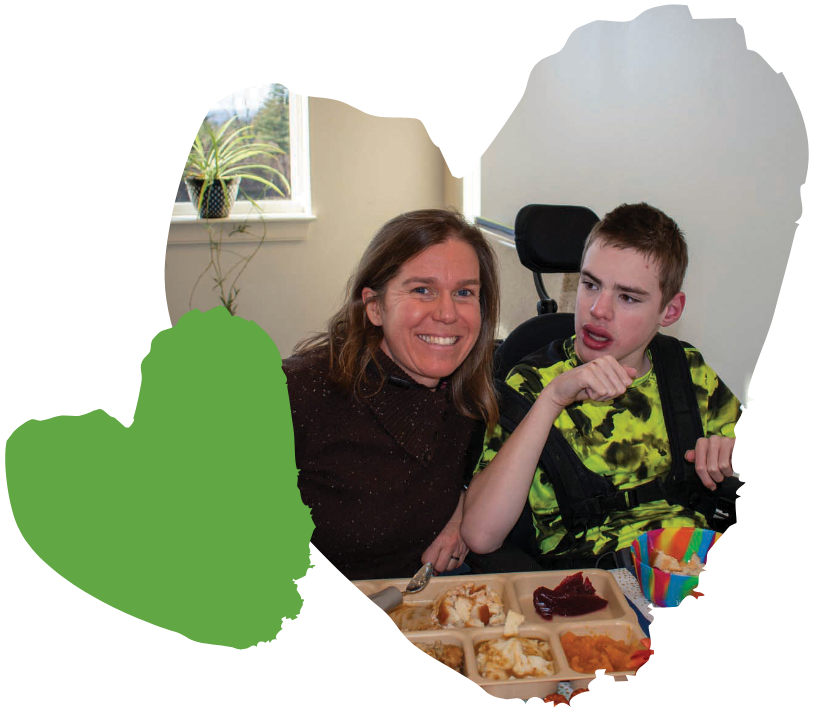 Woman and boy in a wheelchair sitting at a table with food, both smiling. Heart shape overlay and plant in the background.
