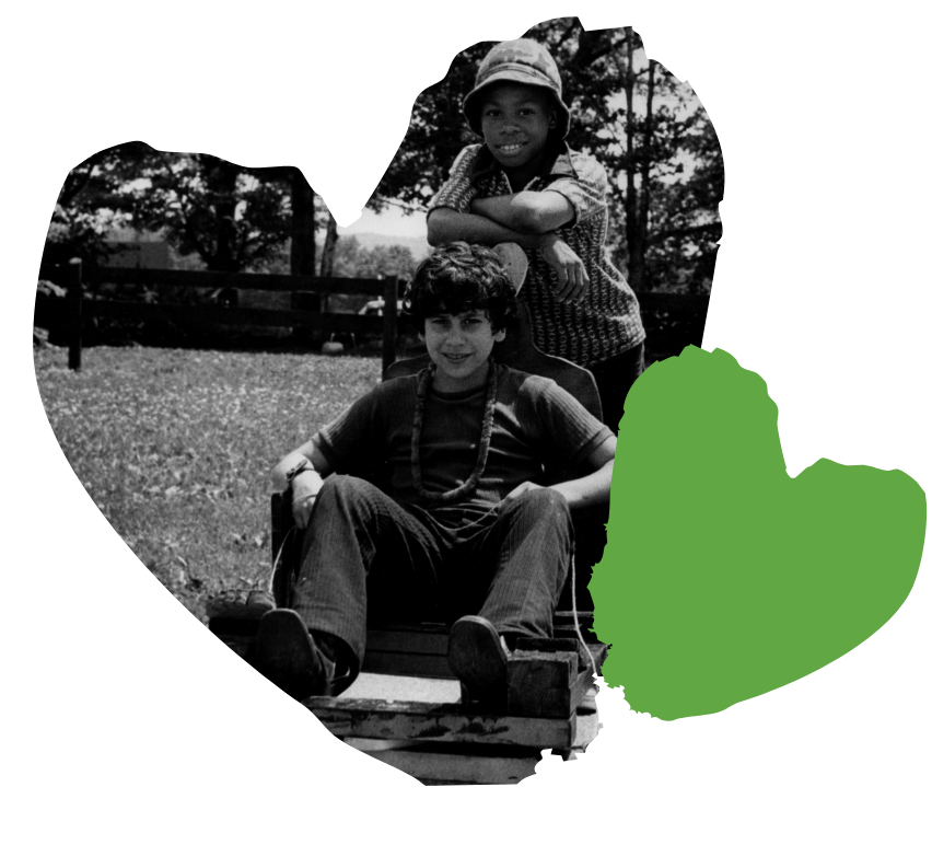 Two children smiling; one sits on a cart, the other leans on it. Theyre outdoors with a green heart shape overlay.