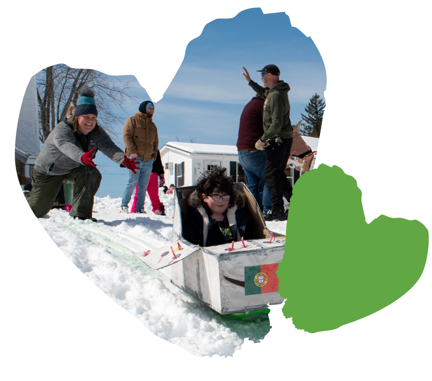 A child rides down a snowy hill in a homemade sled made from cardboard with a Portuguese flag on it. An adult in a blue jacket kneels beside the sled. Other people stand in the background, some wearing coats and hats.