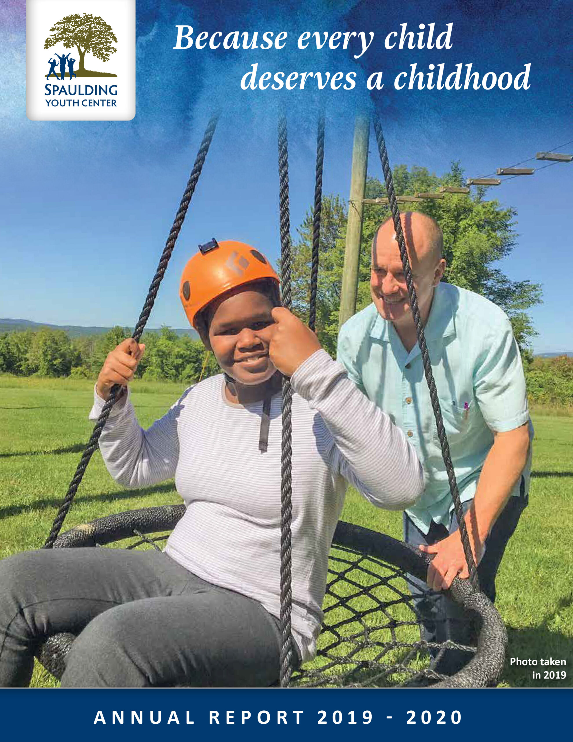 Child on a swing wearing a helmet, assisted by an adult in a grassy park setting.