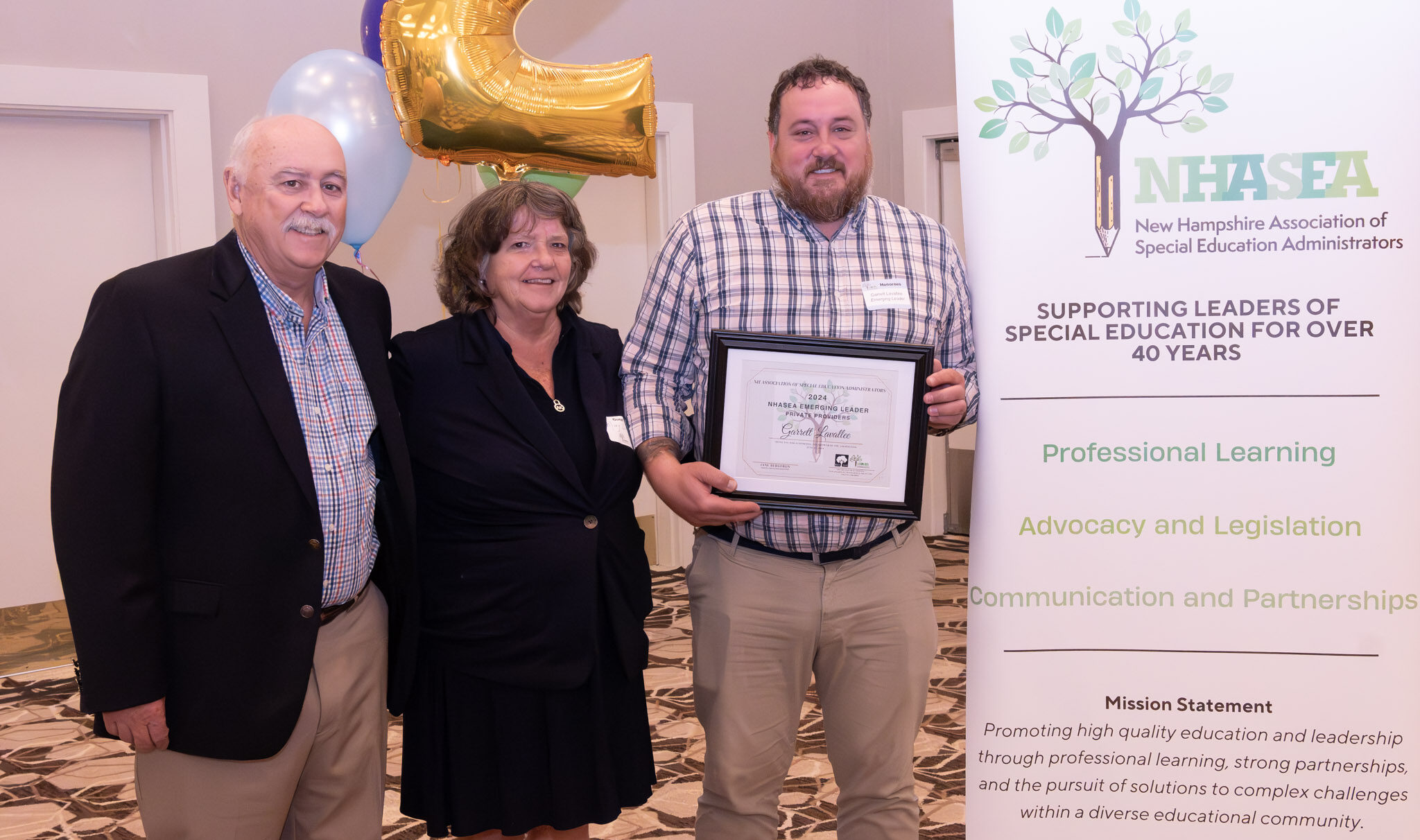 Three people posing with an award certificate beside a NHASEA banner and a gold balloon shaped as the number two.