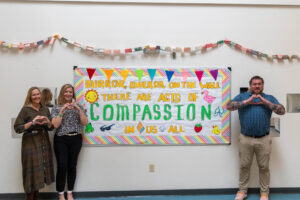 Four people stand in front of a compassion-themed bulletin board, making heart signs with their hands.