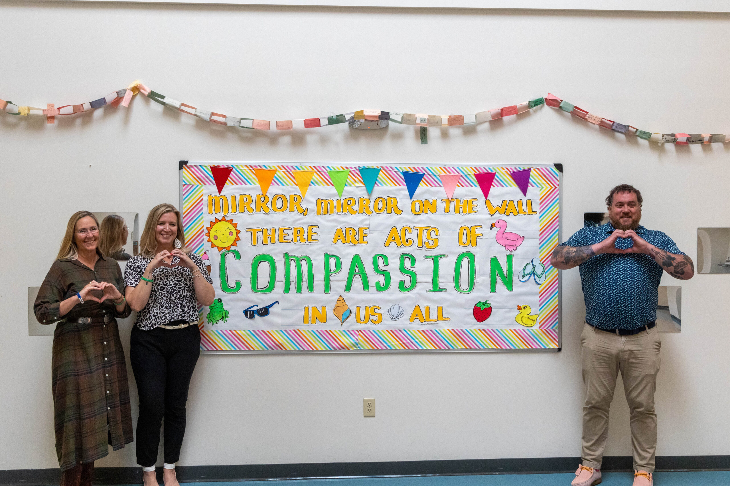 Four people stand in front of a compassion-themed bulletin board, making heart signs with their hands.
