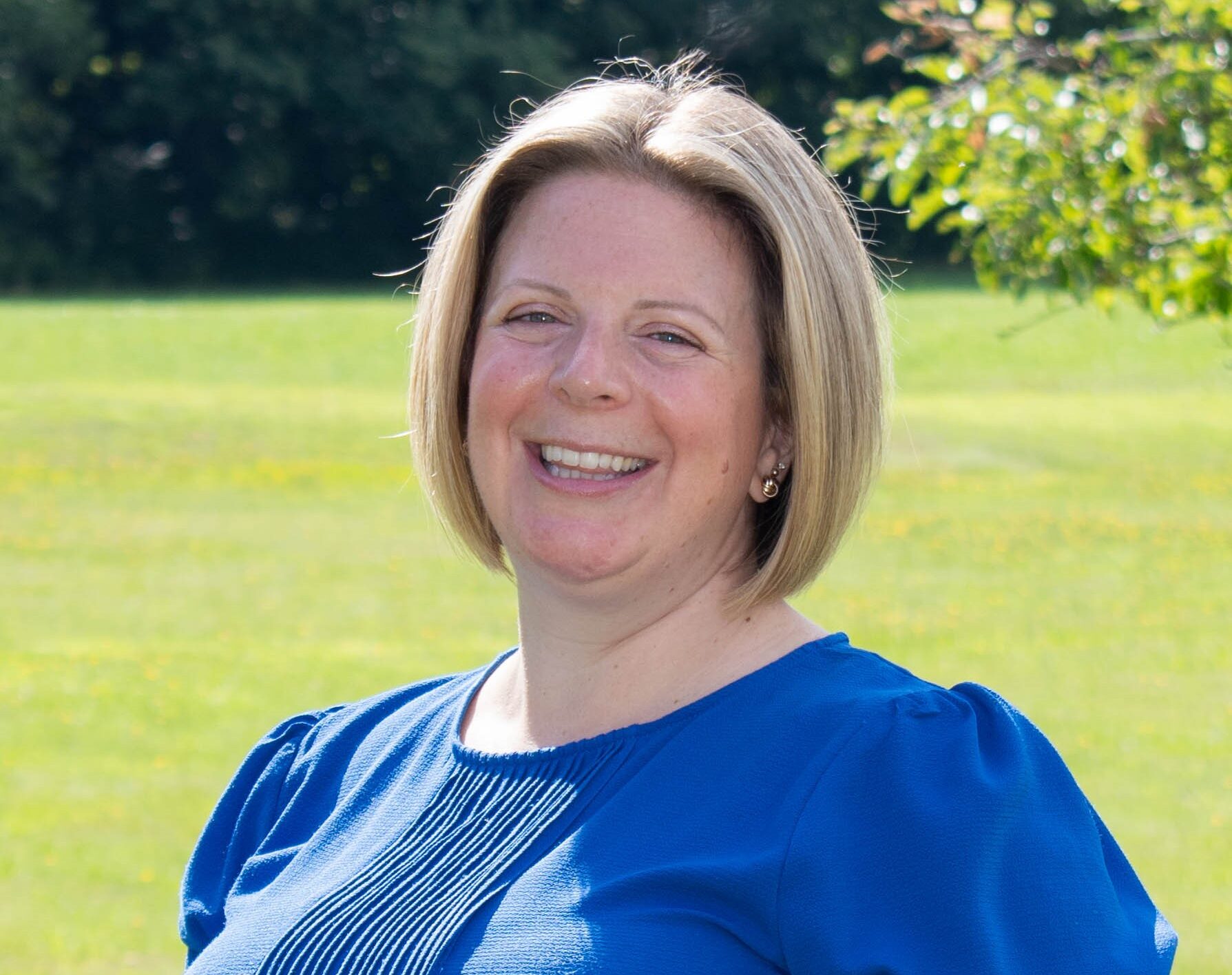Smiling woman in a blue shirt standing outside, green grass and trees in the background.