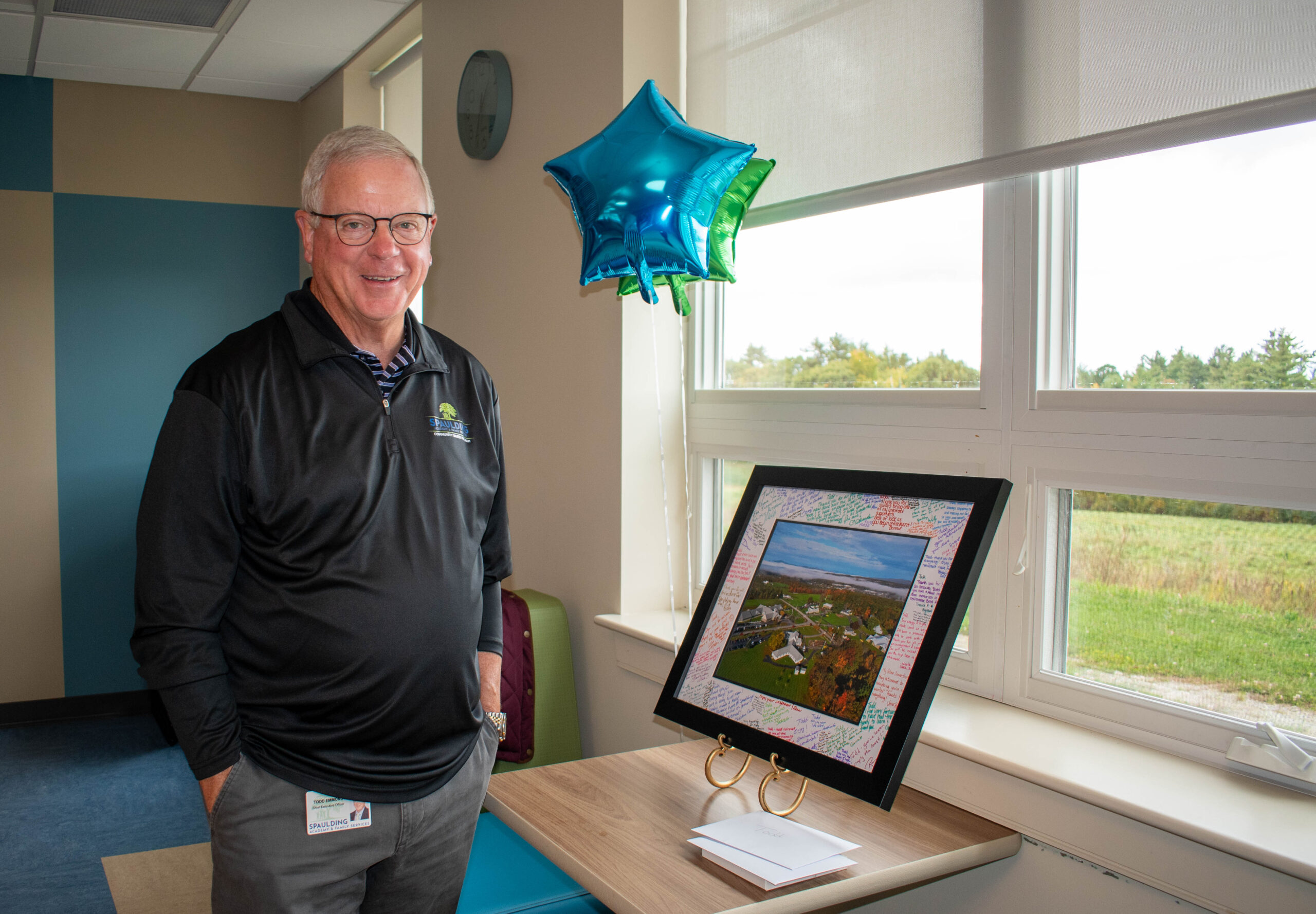 Smiling man stands by a window with star-shaped balloons and a framed photo on a table.