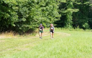 Two people wearing race gear walk on a grassy path beside a forested area on a sunny day.