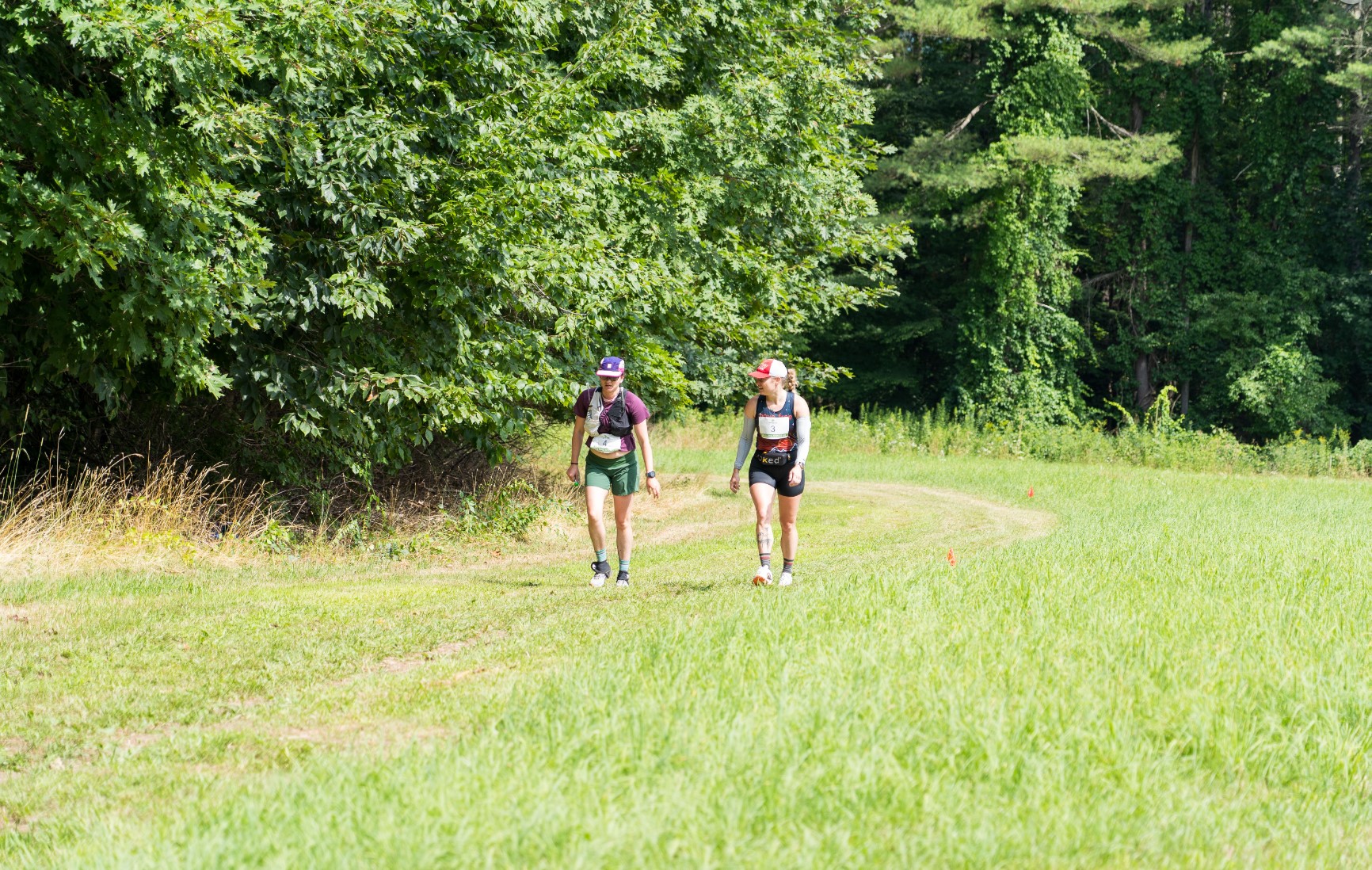 Two people wearing race gear walk on a grassy path beside a forested area on a sunny day.