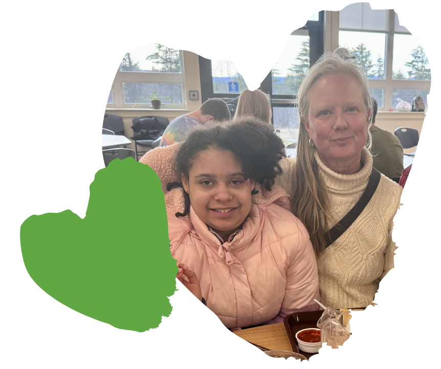 A young girl in a pink jacket sits next to her foster parent, an older woman in a cream sweater. They are inside, surrounded by other people and seated at a table with a lunch tray. A large green heart shape decorates the left side of the image.