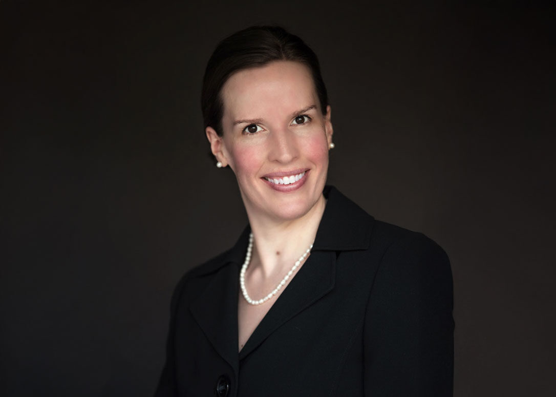 Smiling person in a black suit and pearl necklace against a dark background.