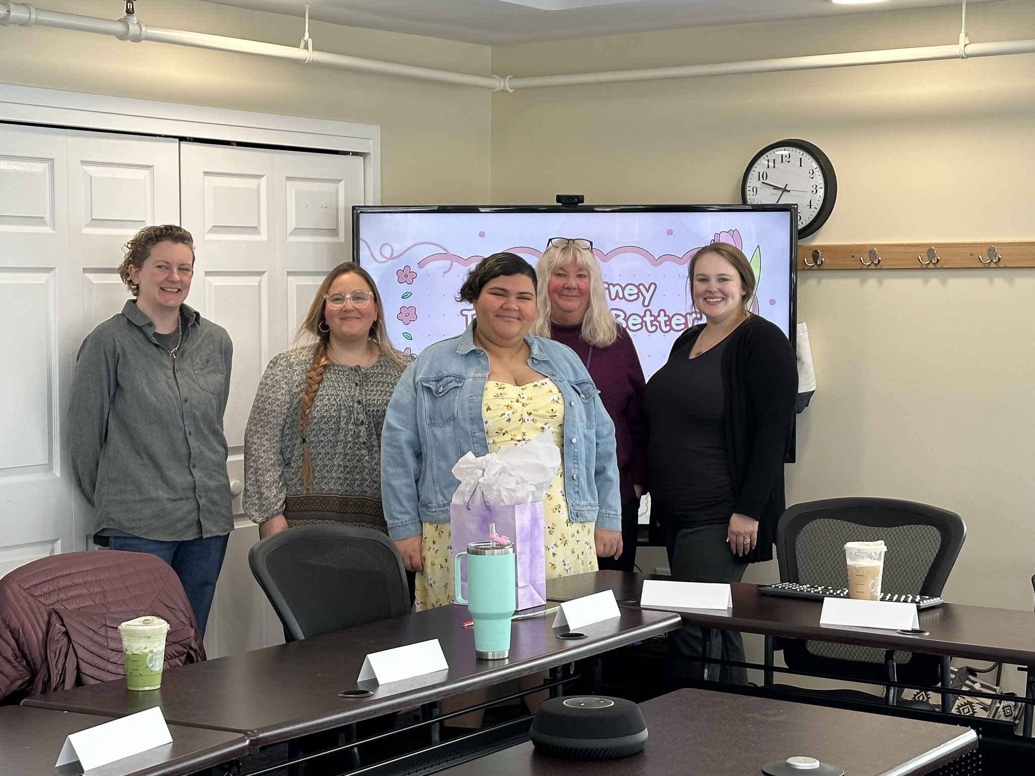 Five people smiling, standing in a conference room with a screen and drinks on the table in front of them.
