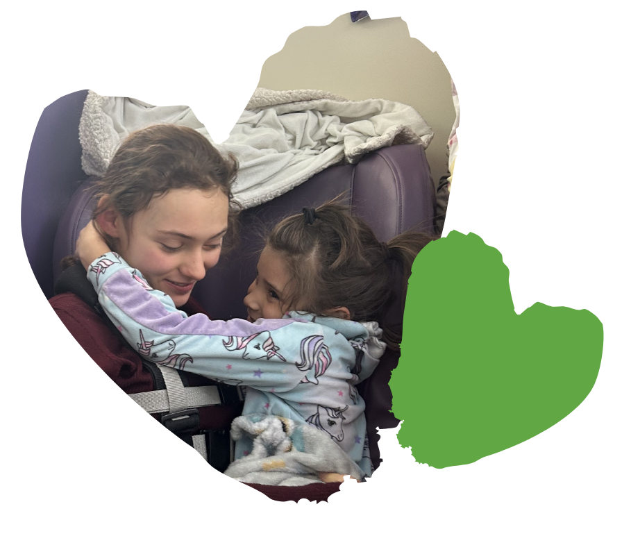 A young woman and a little girl hug and smile at each other, sitting closely on a seat with blankets.
