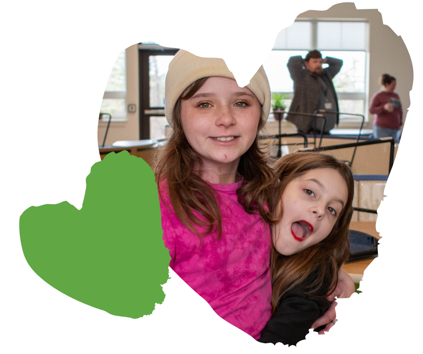 Two smiling girls embrace indoors, with adults and furniture blurred in the background.