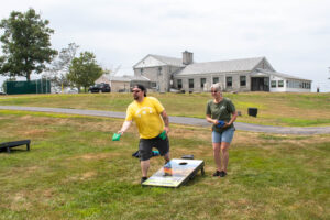 A man and woman play cornhole on a grassy lawn near a large building under a cloudy sky.