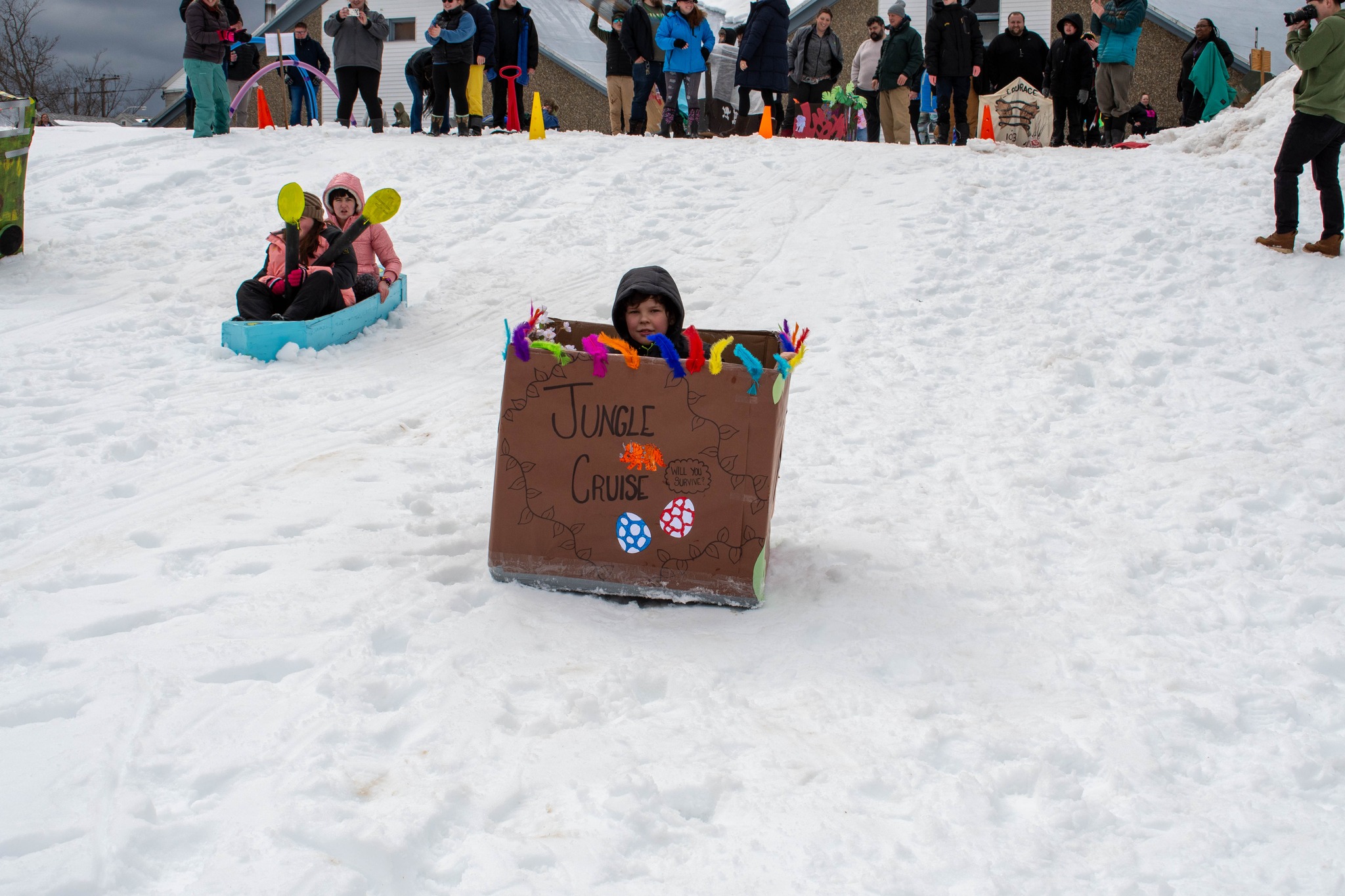 Child sleds down a snowy hill in a decorated cardboard box labeled Jungle Cruise as others watch.