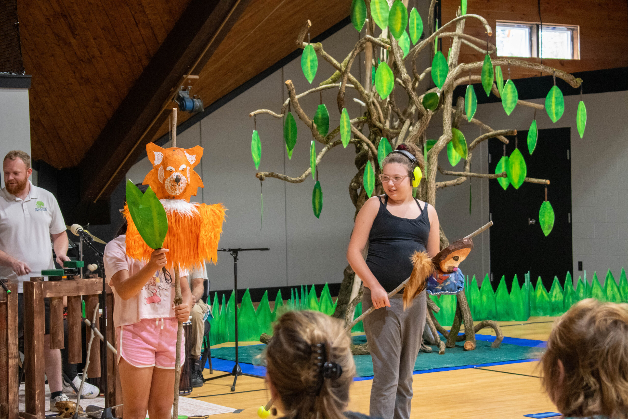 Children perform a play in a gym, one holding a fox mask, with a tree prop and audience watching.