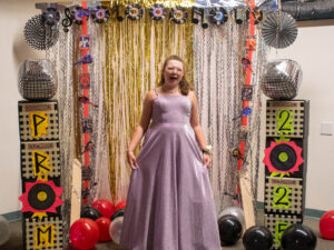 A girl in a sparkly purple dress smiles at prom 2022, standing between colorful decorations and balloons.