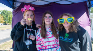 Three people pose under a tent, wearing fun costume glasses and props, smiling at the camera.