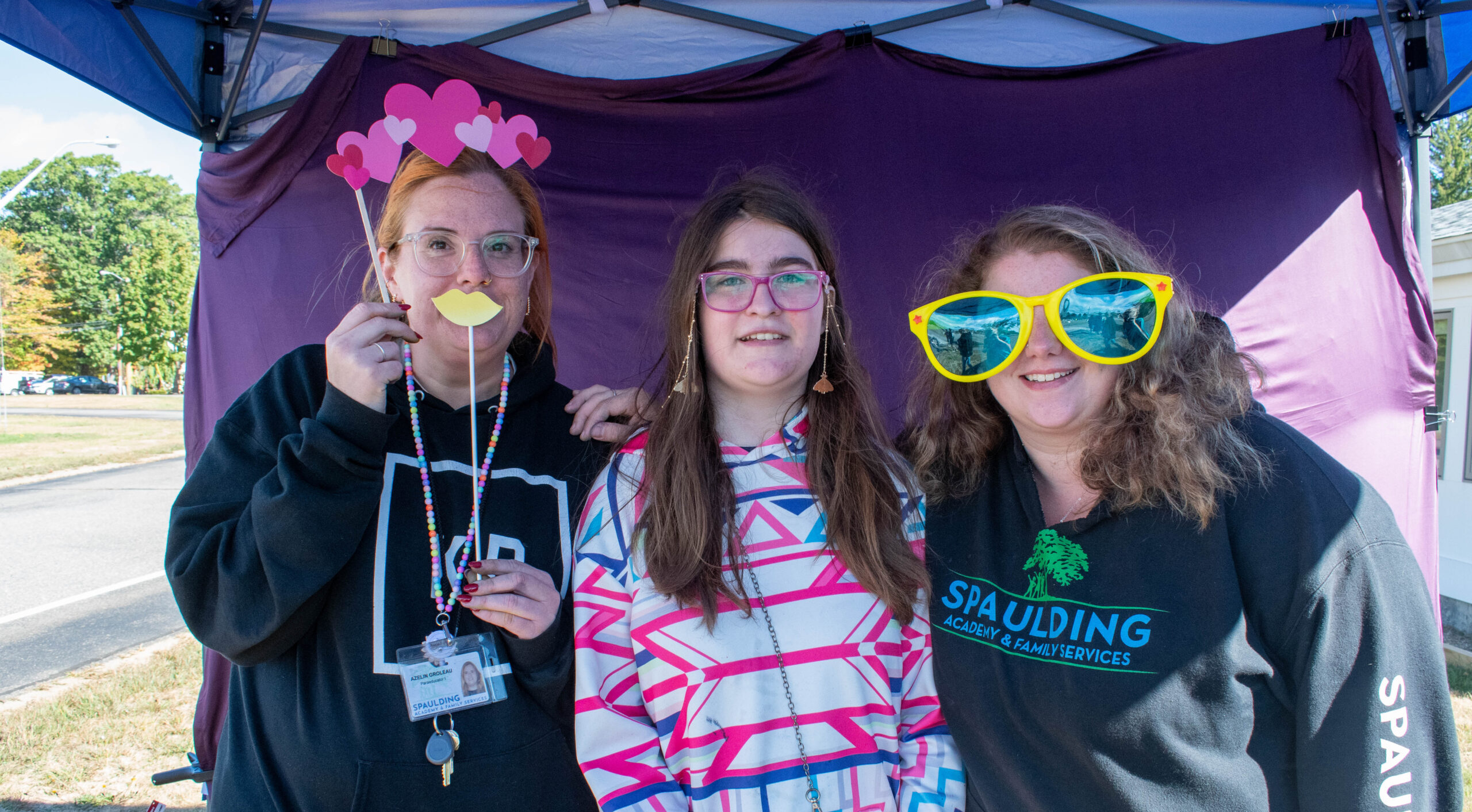 Three people pose under a tent, wearing fun costume glasses and props, smiling at the camera.