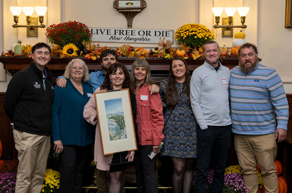 Eight people pose indoors by a decorated mantel with a Live Free or Die New Hampshire sign; one holds a framed picture.
