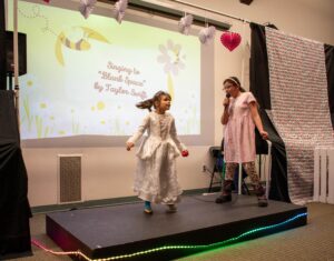 Two young girls perform Blank Space on a small stage, one singing into a mic, the other dancing in a white dress.