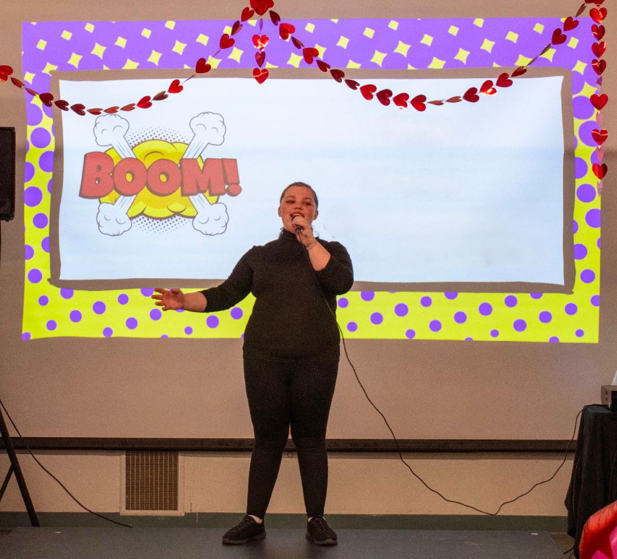 A person sings on stage with a microphone; a colorful BOOM! graphic is projected behind them.
