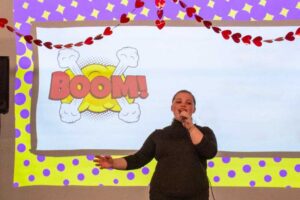 A person sings on stage with a BOOM! comic graphic and heart decorations in the background.