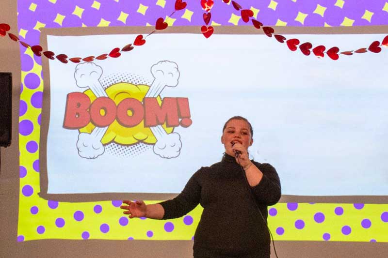 A person sings on stage with a BOOM! comic graphic and heart decorations in the background.