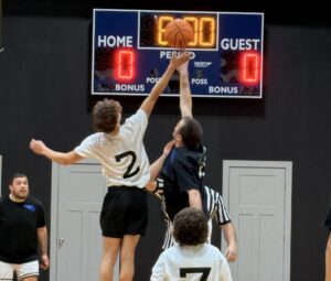 Two youth basketball players jump for the ball at tip-off, while a referee and others watch the action.