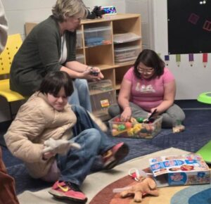 Three people sit on the floor in a playroom, playing with toys and blocks near a green table and yellow chairs.