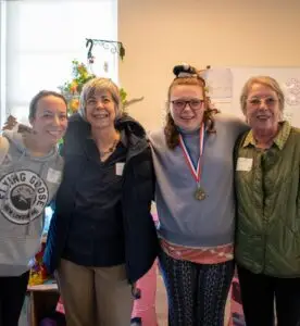 Four smiling women stand together indoors, one wearing a medal around her neck.