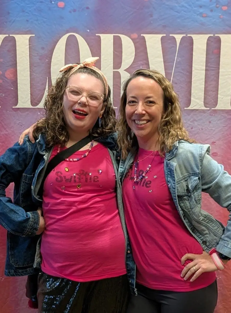 Two smiling women in pink Swiftie shirts pose in front of a pink and purple Taylor Swift backdrop.
