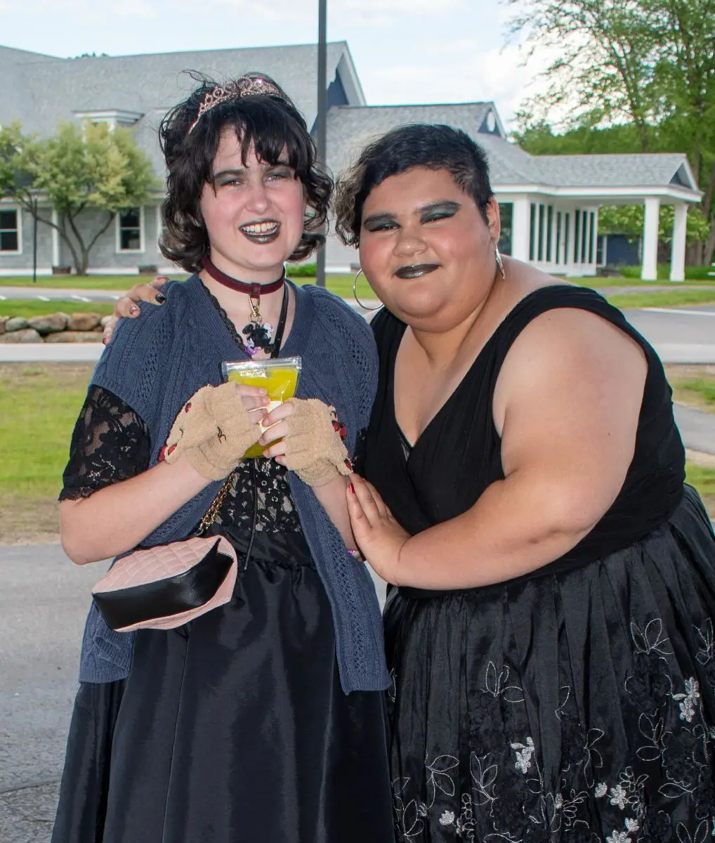 Two people in black dresses and dark makeup smile together outdoors at a formal event.