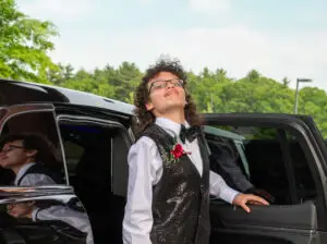 A young person in a tuxedo smiles confidently beside an open car door, with trees in the background.