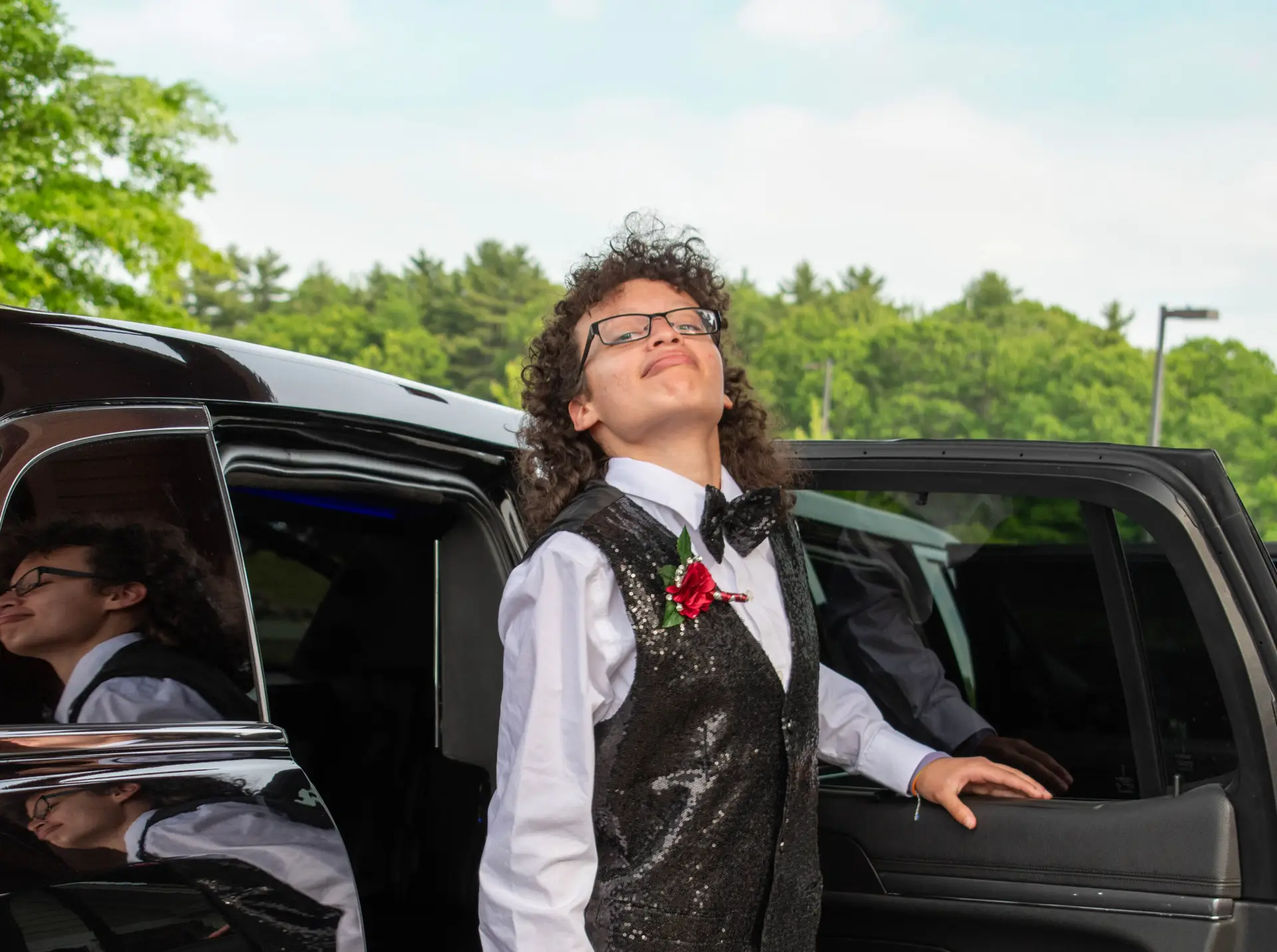 A young person in a tuxedo smiles confidently beside an open car door, with trees in the background.