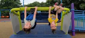 Two smiling kids hang upside down from playground bars, legs hooked, hands gripping, and both wearing glasses.