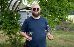 Man with sunglasses holds two small wire tree sculptures outside near a tree and house.