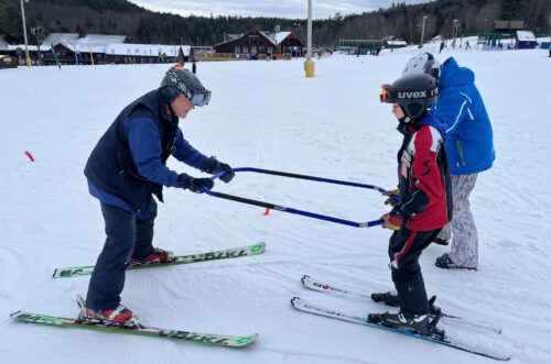 An adult teaching a child to ski using a ski bar on a snowy slope, with another person standing nearby. An adult teaching a child to ski using a ski bar on a snowy slope, with another person standing nearby.