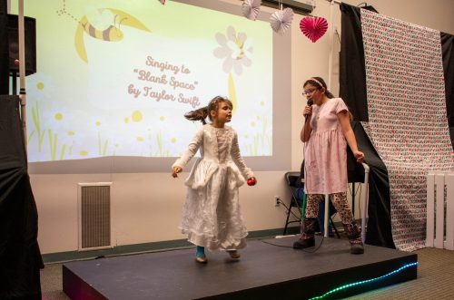 Two young girls perform Blank Space on a small stage, one singing into a mic, the other dancing in a white dress.