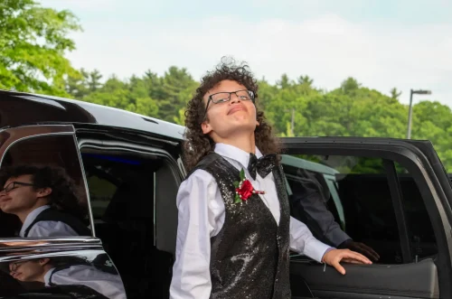 A young person in a tuxedo smiles confidently beside an open car door, with trees in the background.