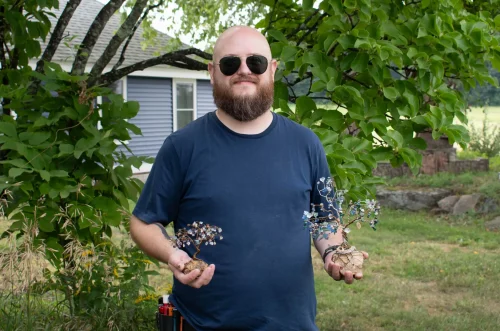 Man with sunglasses holds two small wire tree sculptures outside near a tree and house.
