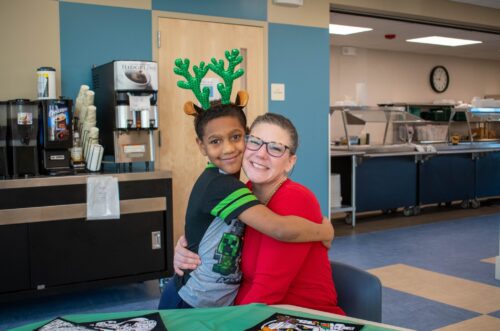Child with reindeer antlers hugs a smiling adult in red, sitting at a table in a cafeteria setting. Child with reindeer antlers hugs a smiling adult in red, sitting at a table in a cafeteria setting.