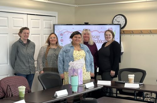 Five people smiling, standing in a conference room with a screen and drinks on the table in front of them.
