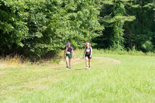Two people wearing race gear walk on a grassy path beside a forested area on a sunny day. Two people wearing race gear walk on a grassy path beside a forested area on a sunny day.