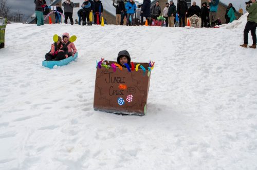 Child sleds down a snowy hill in a decorated cardboard box labeled Jungle Cruise as others watch.