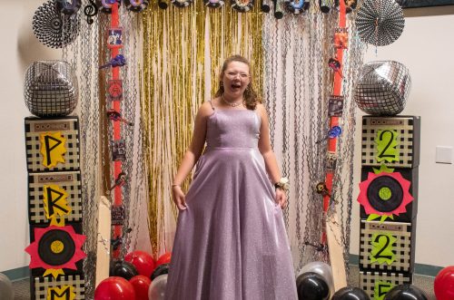 A girl in a sparkly purple dress smiles at prom 2022, standing between colorful decorations and balloons.