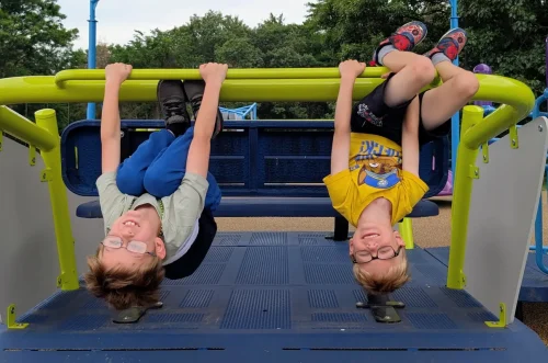 Two smiling kids hang upside down from playground bars, legs hooked, hands gripping, and both wearing glasses.